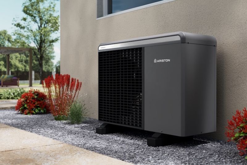 A dark Ariston heat pump beside a house wall and flowering garden plants